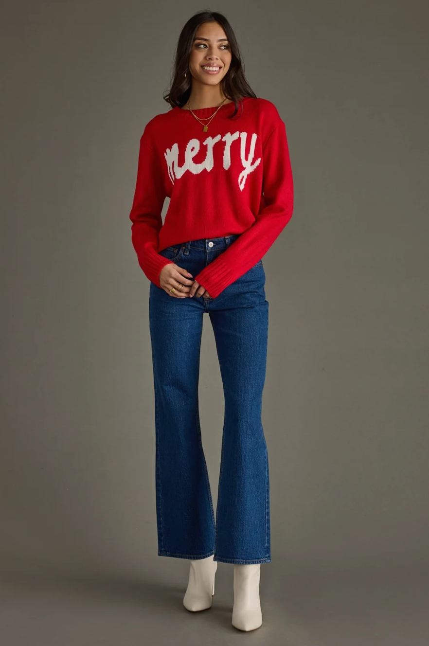 Model wearing a red knit sweater with white Merry text, angled full body view